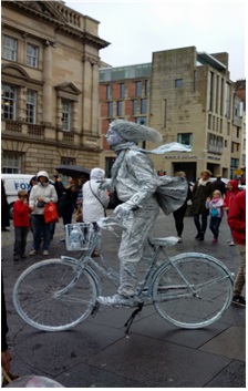 자전거를 탄 해리 포터로 분장하고 서 있는 살아 있는 동상. 머리와 머플러가 바람에 날리는 것처럼 보인다.