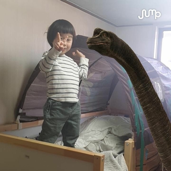 어린이가 침대위에서 목을 빼고 있는 공룡(브라키오사우루스)과 나란히 서서 재밌는 표정을 짓고 있는 모습. 사진=SK텔레콤