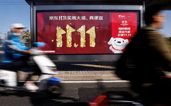 중국 베이징에서 사람들이 전기 스쿠터를 타고 광군제를 홍보하는 징둥닷컴의 광고를 지나가고 있다. 사진=로이터